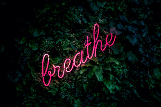 The image is of the word breathe in neon pink against a wall covered in ivy. The intensity of the neon pink illuminates how much of a positive impact breathing has on overall health.