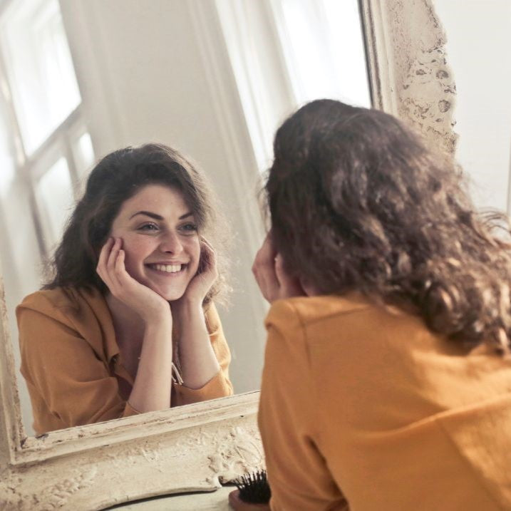 The images shows a young woman staring at herself in a mirror. She is happy and smiling. She is in a large white room with windows.