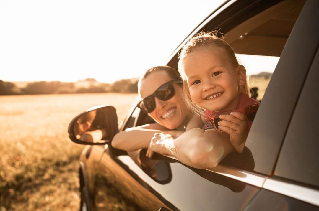 The image shows a mother and daughter looking back from the driver's side of their car with their heads out of the window. They have smiles on their faces.
